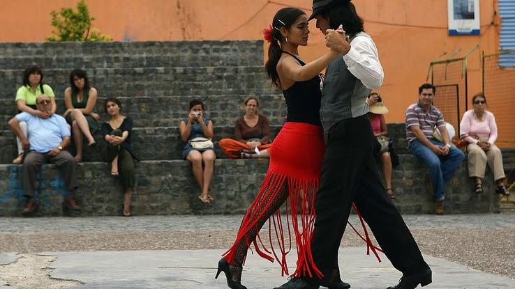 A couple tangoes on the street outside. She is wearing a red skirt with long tassels and he is dressed in black pants and a grey vest. 